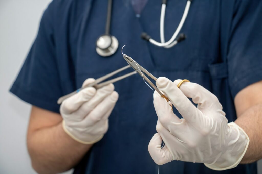 Doctor holds, in hand with disposable glove, forceps and needle holder and suture thread.